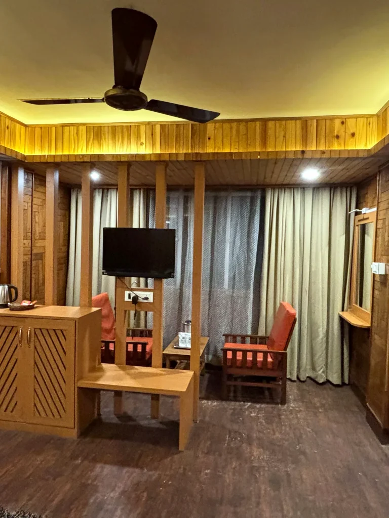 Wood-panelled guest room with a ceiling fan, wall-mounted TV and orange armchairs at Hotel Apsara, Darjeeling